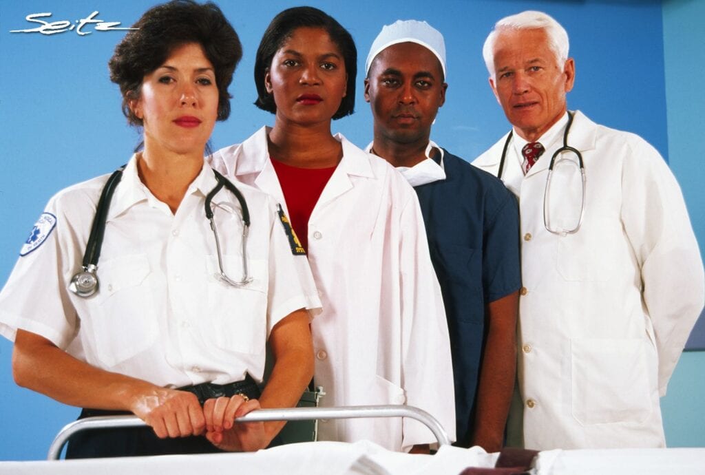 Four diverse health workers, two women and two men, look at the camera over a hospital bed. © Blair Seitz